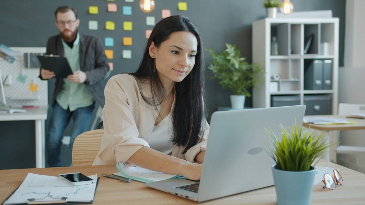 Man is approaching businesswoman with business document talking discussing smiling in office, girl is working with laptop typing. People and communication concept.