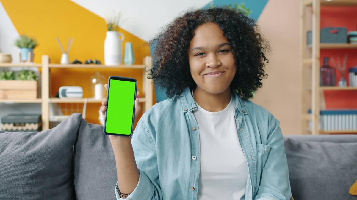 Portrait of attractive African American girl holding green screen chroma key smartphone smiling looking at camera. Technology and modern devices concept.