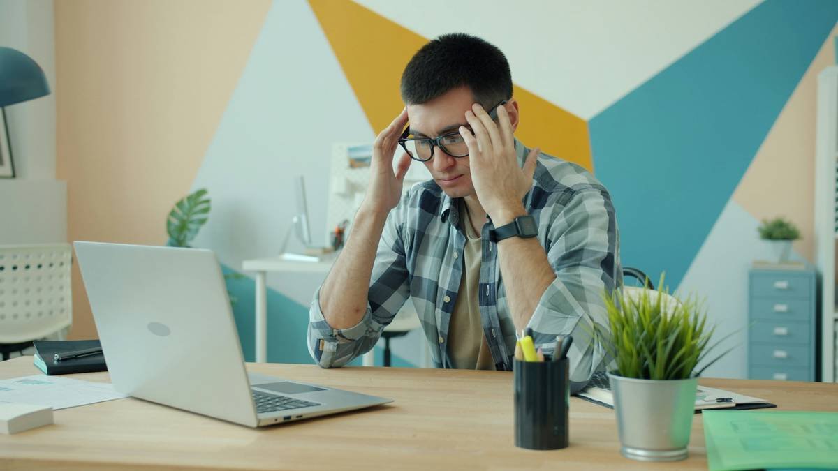 Stressed guy in glasses is working with laptop computer in office suffering from headache overworking touching head massaging temples. Work and health concept.