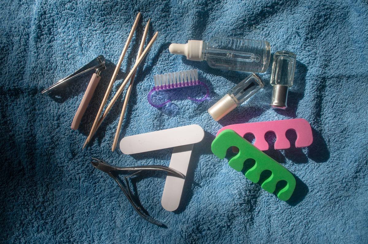 Top view set of pedicure/manicure equipment on a blue towel background on a sunny day. Pedicure kit. Transparent strengthening nail polish, hand serum, clippers, nippers, nail files, nail polisher, nail brush, foam toe separators, orange wood cuticle pusher sticks. Home pedicure tools. Beauty salon, spa, nail bar, nail studio accessories.